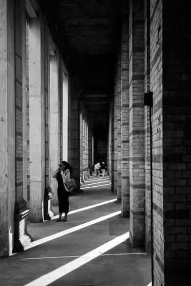 Monochrome photo looking down a tall narrow arcade of square brick columns. The ceiling above is dark, the smooth concrete flow striped diagonally with bright lines of sunlight. In the background, figures walk away from the camera, in the foreground a woman in dark sunglasses stands facing leftwards, right hand up to her mouth, the other holding a phone to her ear.