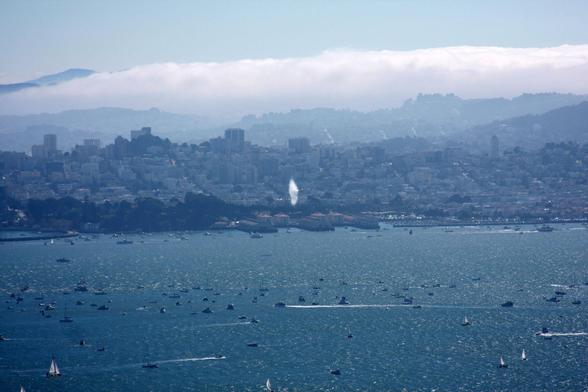 With the San Francisco Bayfront behind it, a Blue Angels jet creates a vapor cone during supersonic flight at low altitude over the water and the many boats below.