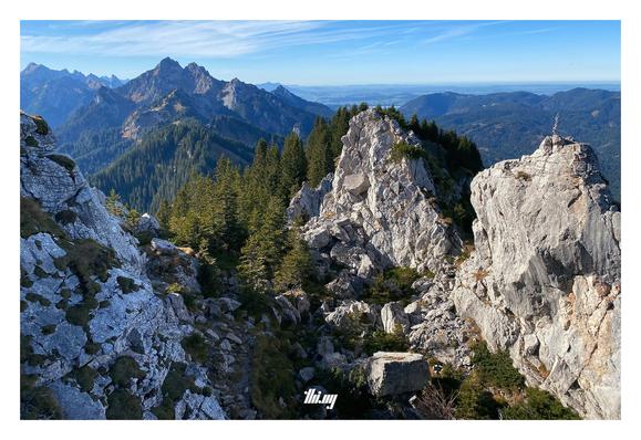 View of white rock cliffs just below a summit in the Ammergauer Alps. Further back, a forested ridge line is leading towards other peaks (e.g. Klammspitze). Pre-alpine low lands in the far, far background. Beautiful sunny day with blue sky.