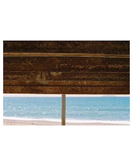 multi layers; above you see rusty horizontal metal structures, then a thin line of sky followed by the ocean and a small stripe of beach. a vertical bar is crossing the sky, ocean and beach line.