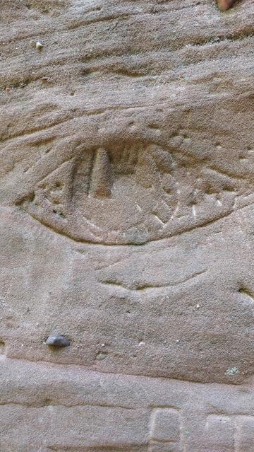 Nahaufnahme eines Felsens aus rötlichem Sandstein. In den Felsen ist ein stilisiertes Auge gemeißelt. Es mutet ein wenig prähistorisch an.

🇬🇧 Close-up of a rock made of reddish sandstone. A stylised eye is carved into the rock. It looks a little prehistoric.