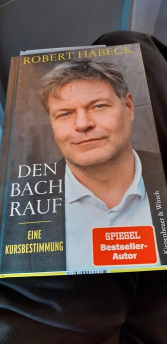 Ein Taschenbuch, das auf meinem Oberschenkel liegt. Das Cover zeigt ein Foto von Robert Habeck, der auch der Autor ist. Der Buchtitel lautet: "Den Bach rauf - Eine Kursbestimmung"