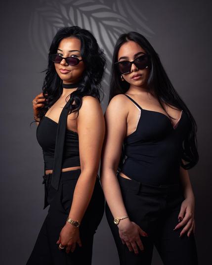 The image shows two individuals standing back-to-back. They are both wearing black outfits and sunglasses. The background is a neutral gray with a leaf pattern shadow. They have long hair and are posing confidently. Valokuvaaja Kuvaajankulma