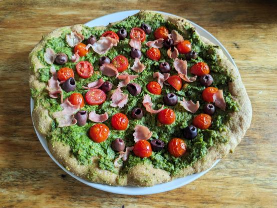 Homemade 12-inch pizza sitting on a plate, featuring whole wheat dough, pesto, tomatoes, olives, and prosciutto. Heavily filtered in the style of Instagram.
