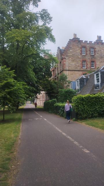 Looking along the path in the Meadows, trees lining either side, now heavy with green, spring foliage, a row of terraced cottages and a Victorian stone tenement on the right