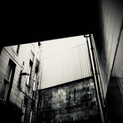 Moody black and white photo with an upward angle perspective; image of the end of an alley where 3 buildings connect. Overhead one building crosses over the alley. The intersection of all of these means there is a shaft of light at the end of the alley which is the subject of this photo. Pipes, windows, brick, metal siding are all visible on the building façades. The image has a strong linear geometry.