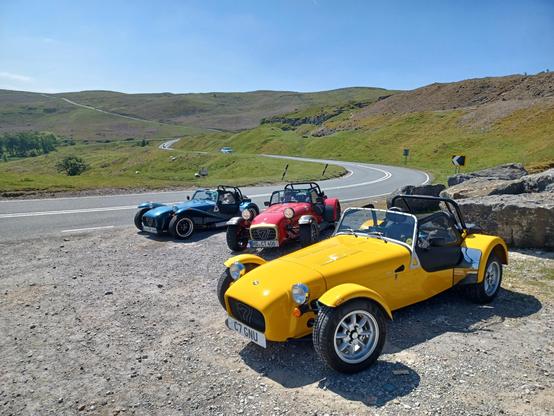 Yellow, red and blue Caterham 7s parked in a layby on the Black Mountain Road in the Brecon Beacons National Park, Wales. The twisty road is in the background.