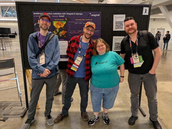 Four Wagtail community members standing next to each other in front of the Wagtail poster in the exhibit hall at PyConUS.