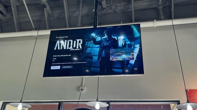 A public video screen hanging from and industrial-type ceiling. The display is showing the start screen for Andor Season 1 Episode 2 That Would Be Me