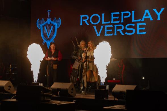 Color photo of three persons on stage at RPV 2025, among them Maul Cosplay in his Geralt of Rivia costume. There are pyrotechnical effects on stage and a projection says "ROLEPLAY VERSE" and shows the RPV Logo.