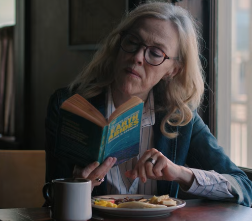 a picture taken from "the last of us" s02e06 showing a women with semi long blond airs and glasses at a table reading a used book titled "EARTH ABIDES" in yellow over blue background cover