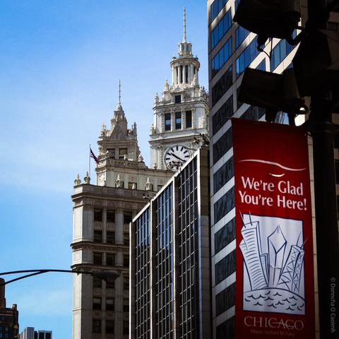 A street scene in downtown Chicago featuring the ornate clock tower of the Wrigley Building under a clear blue sky, with a red banner on a lamppost in the foreground that reads: "We're Glad You're Here! CHICAGO Mayor Richard M. Daley" alongside a simple line drawing of skyscrapers and waves.