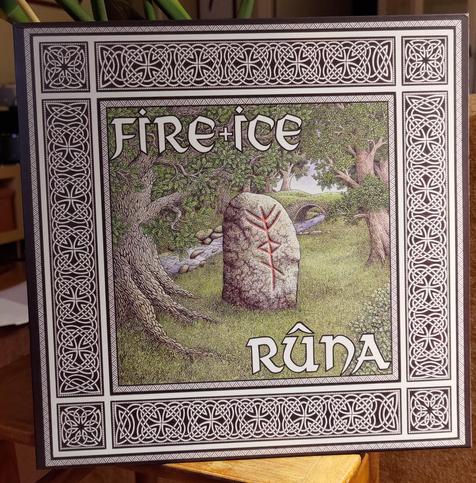 LP cover standing on a small table with a plant behind it.

A drawing of a rune stone in the wood, with green grass and some trees with green leaves. A thick frame with a black and white pattern.

Top left on the drawing: "Fire + Ice" in large white letters.
Bottom right: "Rûna"