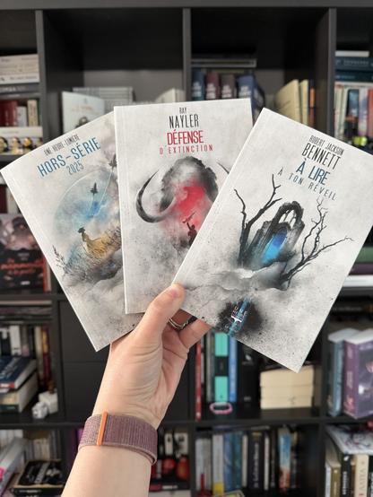 Livre pris en photo devant une bibliothèque. 
La couverture d’A lire à ton réveil montre un morceau de cathédrale en ruine avec une porte bleue et deux arbres morts sur les côtés.
La couverture de Défense d’extinction montre un mammouth avec devant une personne levant une arme d’assaut. 
La couverture du Hors-série UHL 2025 montre une femme dans un champ regardant dans le ciel trois vaisseaux qui s’envolent devant une planète.