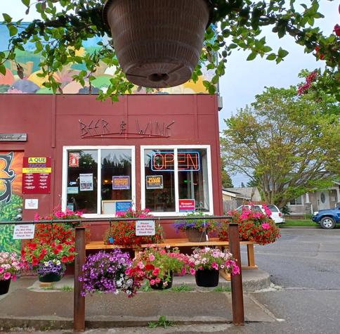The storefront of a small, flat topped square building with colorful flowering baskets hanging from the handrail leading to the entrance. In the foreground at the top of the photo is the bottom of a hanging plant with its greenery spilling out over the sides of the pot. In the window of the storefront are signs in support of the local nurses union.