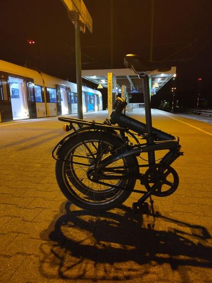 Ein Faltrad steht zusammengeklappt auf einem S-Bahn Steig.
Links hält gerade eine Bahn mit offenen Türen.

Es ist Nacht und die Beleuchtung ist bahntypisch schummrig-honiggelb