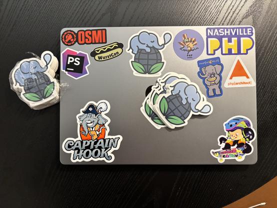 A laptop with stickers on it. The featured sticker is the one I had made for REST Certain; it is of a cute elephant sleeping atop a globe. A stack of these stickers is fanned out on top of the laptop, and another stack still in plastic wrap is off to the side. The laptop additionally has stickers for OSMI, PhpStorm, WurstCon, Captain Hook, Longhorn PHP 2023, Nashville PHP, PHP World 2019, PHP Architect, and GitHub.