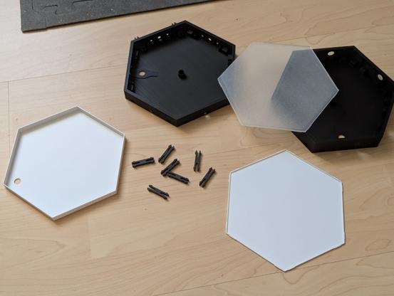 Auf einem hellbraunen Fußboden liegen mehrere mit einem 3D-Drucker gedruckten Hexagonalen Elemente in weiß und Schwarz, so wie ein paar Steckverbinder dafür.