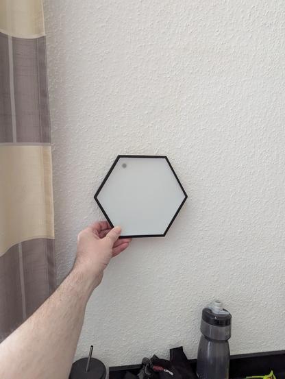 Ein Arm und eine Hand, die ein Hexagonales Element vor eine weiße Wand hält, um zu schauen, wie es optisch aussieht.
