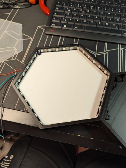 Ein schwarzes, hexagonales Element, welches im Innern mit LEDs beklebt und der Boden mit einem weißen Plastikelement abgedeckt wurde. Links daneben liegt eine weitere transparente Abdeckung, die später oben drauf kommt, am rechten Rand ein weiteres Element, welche mit Steckverbindern mit dem ersten Element verbunden wurde.