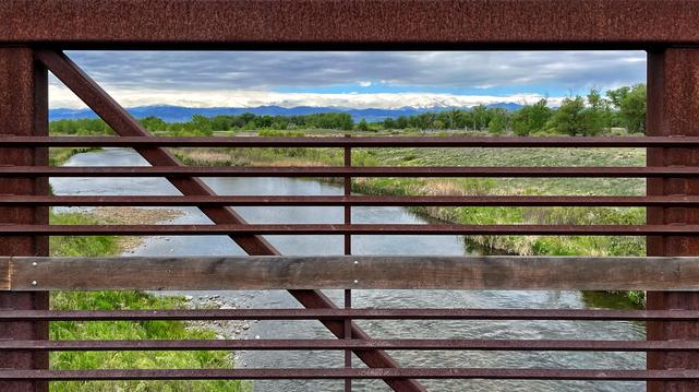 Peering through the horizontal, rusty metal rails on a pedestrian bridge across a large flowing creek, with grasses, shrubs, and trees along the banks of the creek on either side, and a line of foothills and mountains in the far distance, under mostly cloudy skies with a few peeks of blue.