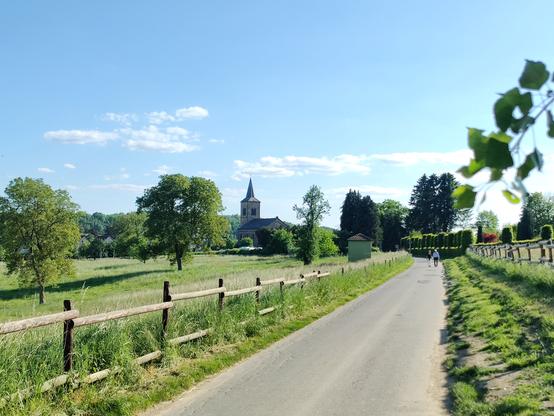 Eine schmale Straße, links und rechts davon Holzzäune und grüne Wiesen. Im Hintergrund ist din Kirchturm zwischen den Bäumen zu sehen.