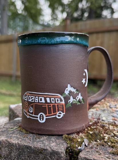 Brown clay ceramic mug with a camping van  and mountains stamped into the clay and highlighted with orange and white underglaze. Mountains are the moon and 2 stars are in the background highlighted by green and white underglaze.