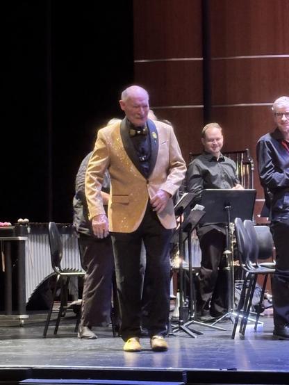 A conductor wearing a sparkly gold jacket and bow tie walks on stage, with band members and musical instruments in the background.