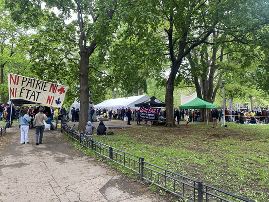 A hundred or more people gathered in a park, with some large tents set up, a line for food, and a banner reading Ni Patrie Ni État Ni Québec Ni Canada