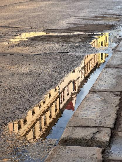 the roofline of a row of apartment buildings, coloured from cream to pale yellow, reflecting in a long puddle in the gutter of a street