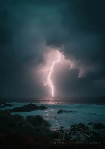 A powerful lightning bolt strikes the ocean beyond the rugged coastline of Phillip Island, casting a brilliant white glow that pierces the dark storm clouds above. Jagged rocks line the shore in the foreground as waves crash beneath the glowing sky. The moody teal and indigo tones create a dramatic contrast with the electric burst of light.