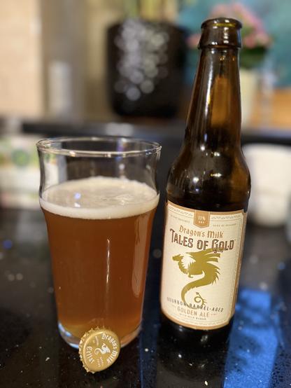 A pint glass of golden ale next to a brown bottle with a gold and tan label. A gold dragon 🐉 in silhouette is in the center.
