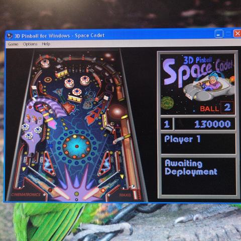 The image shows a screenshot of the "3D Pinball for Windows - Space Cadet" game. The main part of the image displays the pinball table, which is colorful and features a space theme with various pinball elements such as ramps, targets, and flippers. The table is labeled "CINEMATRONICS" and "MAXIS." On the right side, there is a smaller window with the game's title "3D Pinball Space Cadet" and a cartoon character in a spaceship. The score for Player 1 is 130,000, and it is labeled as "Ball 2." The text "Awaiting Deployment" is also visible. The game interface is set against a background that includes a green parrot and a rocky surface, adding a natural element to the scene.