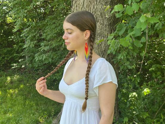 Yocheved standing by a tree and holding one of her braids. She's wearing a white flowy top and a pair of Special Edition Hvresse (Moon) Rainbow Pride Fringe Earrings.