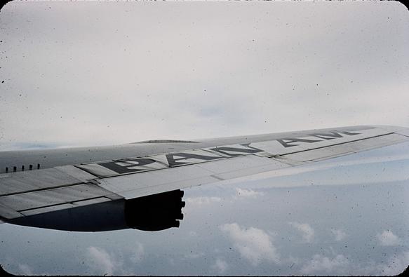 The starboard wing of a Pan Am plane, the words Pan Am painted on the wing. It's cloudy below.
