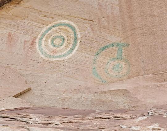 A color landscape photo of two pictographs painted on beige colored sandstone rock. The one of the left is a circle about a foot in diameter, that looks like a bullseye. It has a white center and a thick green line circumscribed around the center. A similar width white line is circumscribed around the green one. Then there is a space about four times the width of the lines. After the wide blank space there's a white line, then a bit thicker green line, and finally a white outer line. To the right of the circle is a second pictograph. It is green. A green semicircle, the same outer size as the pictograph on the left, is on the left side. A bar extends down from the top part of the semicircle to a smaller green circle. The center of the small green circle has a green dot.
