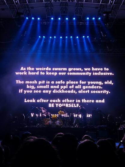 Photo of an elaborate stage with a very large rear projection display behind the band (King Gizzard and the Lizard Wizard) who have yet to take the stage. The display shows a simple text message:

As the weirdo swarm grows, we have to work hard to keep our community inclusive.
The mosh pit is a safe place for young, old, big, small and ppl of all genders.
If you see any dickheads, alert security.
Look after each other in there and BE YOURSELF
[illegible line]