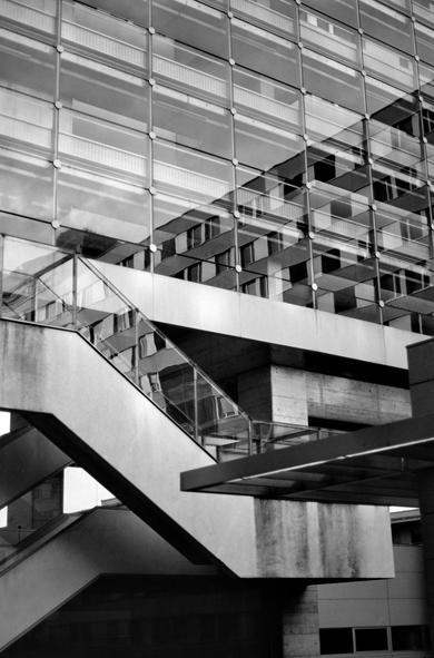 The black and white image shows a modern building with a large glass facade reflecting another structure, creating a layered visual effect. A concrete staircase with glass railings diagonally crosses the frame, adding a dynamic architectural element. The overall scene emphasizes geometric shapes and urban symmetry.