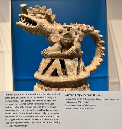 photo of the incense burner (closeup of caiman top) and gallery label “For many cultures of Latin America, the Earth is visualized as the back of a giant caiman or crocodile floating in a primordial sea. Such a large reptile rears its head from the top of this incense burner. Incensarios were used in rituals to burn the resin of the copal tree. Its smoke was thought to protect against respiratory illnesses and headaches. In Central America, burners like this are often found broken in shrines on the slopes of a volcano in Lake Nicaragua. Their smoke would have imitated the volcano, and burning incense was likely a form of care and offering for the living landscape. Caiman Effigy Incense Burner Unidentified artists), Guanacaste-Nicoya culture, Costa Rica or Nicaragua, 500-1350 CE Earthenware, traces of white ground Gift of John G. Bourne, 2009, acc. no. 2009.20.45”