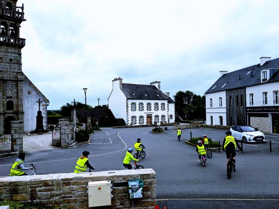 6 enfants et 2 adultes à vélo, en tenue fluorescente et casqués, déambulent dans un le bourg d'une commune devant le parvis d'une église et autre commerce