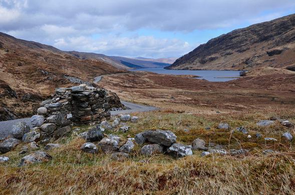 The remains of Tigh nan Saighdearan. The image shows a view down a broad glen with a loch beginning halfway along it and continuing into the distance. There’s a gravel track coming in on the left and continuing along the hillside. In the foreground are partial remains of stone walls. The light is grey.