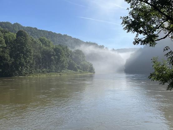 An einem sonnigen Morgen fließt ein dunkelgrüner Fluss zwischen bewaldeten Hügeln in ein schmales Tal, in welchem noch der Nebel steht.
