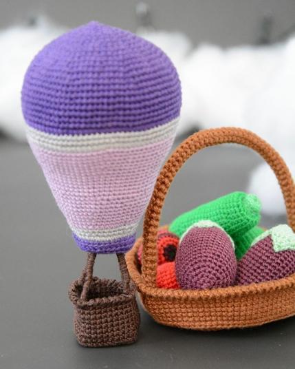 Gros plan sur une montgolfière crochetée aux tons violets, posée à côté d'un panier rempli de légumes crochetés. La nacelle et le panier sont bordés avec un rang de mailles écrevisse, visible en relief.