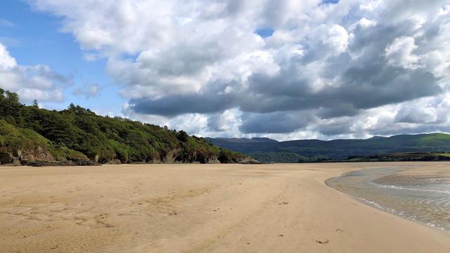 This picture captures a stunning coastal landscape in Gwynedd, Wales, near Portmeirion. The foreground features a serene expanse of shallow water gently meeting sandy tidal flats. On the left, lush green hills with dense vegetation slope down toward the water’s edge, while the right side showcases rolling hills and distant mountains covered in varying shades of green. The sky is dramatic, filled with large, fluffy clouds casting shadows over the landscape, creating a beautiful contrast between light and dark areas. The peaceful scenery exudes a natural, untouched charm typical of the Welsh coast.