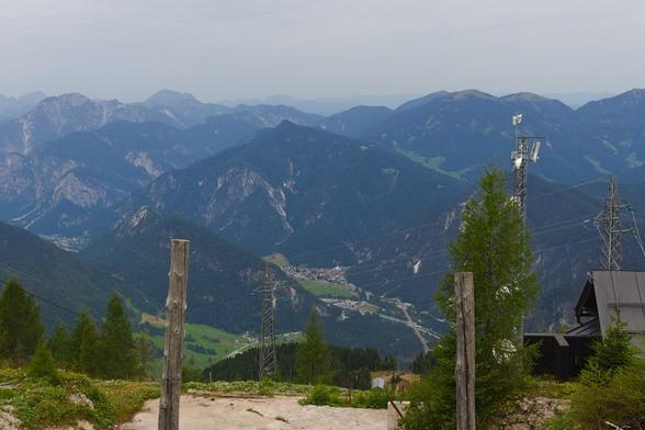 This photo captures a breathtaking mountain valley view from Monte Lussari. The scene looks westward over steep, forested slopes and rugged grey peaks under an overcast sky. In the distance, layers of blue-grey mountains recede into soft mist. A small village lies nestled in the valley far below, its red roofs providing a warm contrast to the cool tones. The foreground includes a few tree trunks, grassy patches, and utility towers with cables cutting across the view. A radio tower and rustic rooftops appear on the right. The overall mood is serene and vast, offering a sense of height and isolation.