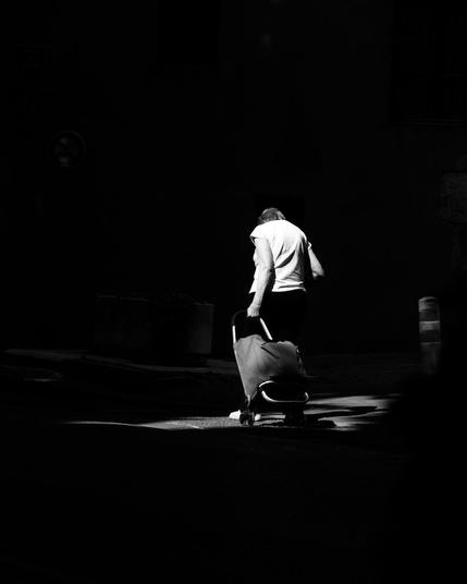Vieille dame trainant son chariot à provisions dans un rai de lumière. Noir et Blanc