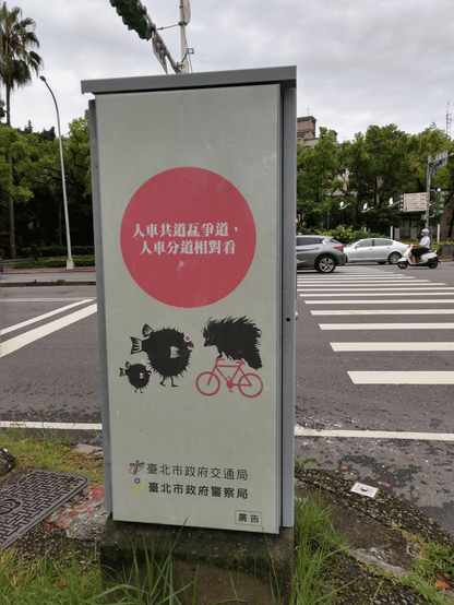 Sing near a road and pedestrian crossing, reminding cyclists and pedestrians to watch out and take extra care since they share the same space here. Cyclists are represented graphically by a hedgehog on a bike in full speed and mean eyes, the pedestrians are spiked pufferfish with widened, scared eyes. All writing is in Chinese characters. The road is several lanes wide for each direction, the traffic light shows red towards the camera, a few cars are visible. Sky is overcast