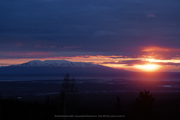 The bright orange sun almost touches the horizon on the far right side of the frame.  The snow-capped silhouette of Sleeping Lady is backlit by a light band of sunset light.  Dark clouds at the top of the frame.