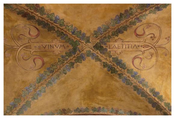 This is a painted ceiling with warm ochre tones and decorative grapevines forming an X shape. Each diagonal line is made of grape leaves and clusters of purple and green grapes, symbolizing abundance. In the horizontal arms of the X are two Latin words in ornate frames: “VINVM” on the left and “LAETITIA” on the right, meaning “Wine” and “Joy.” Elegant swirls and floral flourishes surround the words, and the symmetrical design evokes classical and religious frescoes. The overall feel is celebratory, emphasising wine as a source of happiness and festivity in a traditional, richly detailed setting.