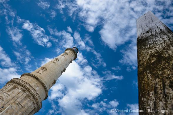 Le phare de Cordouan photographié en contre plongée sur un ciel bleu animé par des nuages blancs. A sa droite, un pieu de bois marqué par les marées, pointe lui aussi vers le ciel.